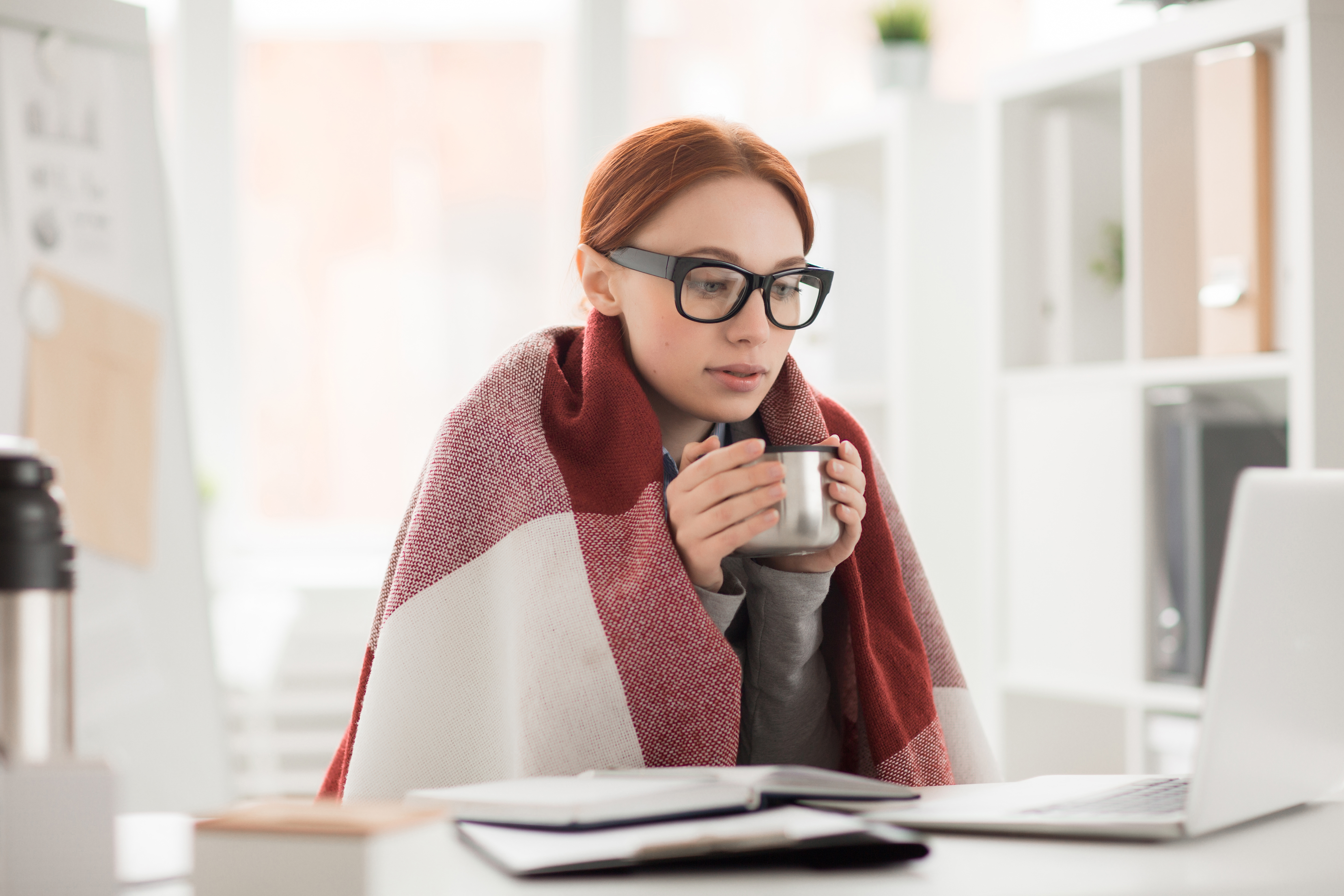 Mujer joven con gafas, sentada frente a un escritorio mirando concentrada a la pantalla de un portatil mientras sostiene una taza metálica entre ambas manos. Tiene una manta cubriendo sus hombros.